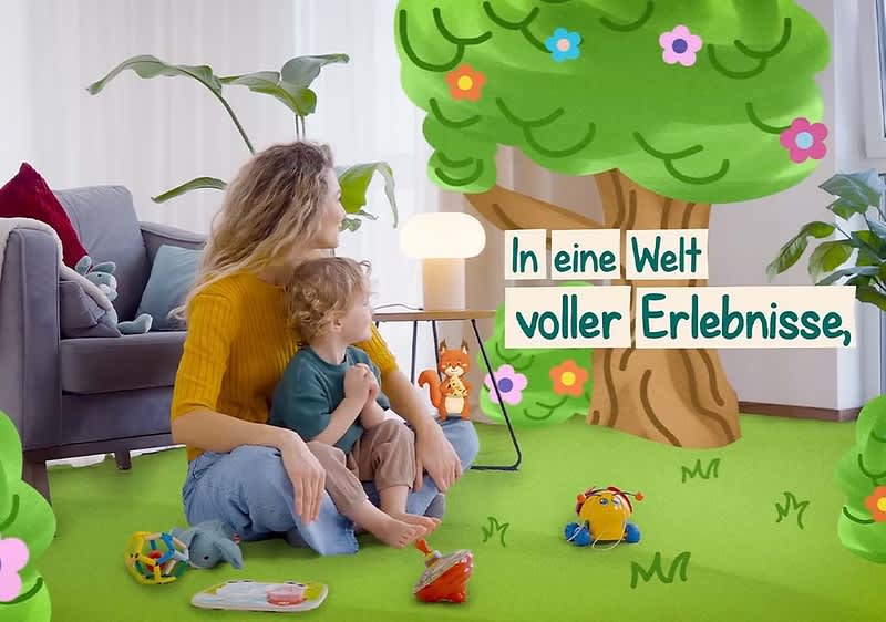 Mutter und Kleinkind sitzen auf grünem Teppich vor einem Wohnzimmerfenster, die Mutter hält das Kind auf ihrem Schoß, beide schauen nach rechts, umgeben von Spielzeug; im Vordergrund und an der Wand über einem Baum-Illustration der Text „In eine Welt voller Erlebnisse“ ohne Satzendezeichen