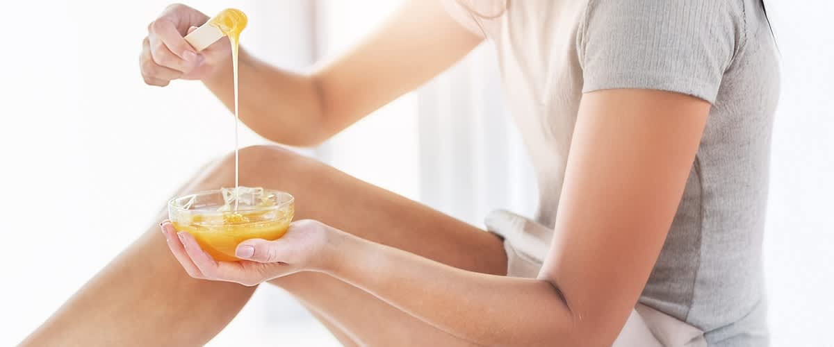Hände einer sitzenden Person halten eine kleine Glasschale mit warmem, goldgelbem Zuckerwachs, mit einem Holzspatel wird das Wachs herausgezogen und bildet einen langen Faden, im Hintergrund ist ein angewinkeltes Bein sichtbar