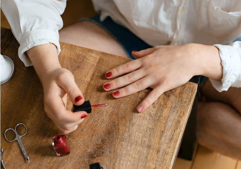 Eine Person lackiert mit der linken Hand die Fingernägel der rechten Hand mit rotem Nagellack, auf dem hölzernen Tisch neben einer offenen Nagellackflasche und einer Nagelschere liegend, die Person trägt ein weißes Oberteil und dunkle Shorts