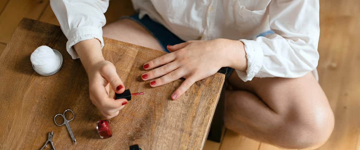 Eine Person lackiert mit der linken Hand die Fingernägel der rechten Hand mit rotem Nagellack, auf dem hölzernen Tisch neben einer offenen Nagellackflasche und einer Nagelschere liegend, die Person trägt ein weißes Oberteil und dunkle Shorts