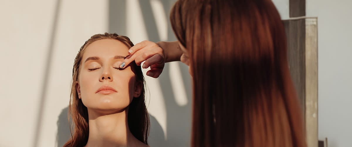 Frau sitzt frontal im Bild, Haare sind nass und werden von einer zweiten Person mit den Fingern am Haaransatz zurechtgestrichen, heller Hintergrund mit Schattenmustern
