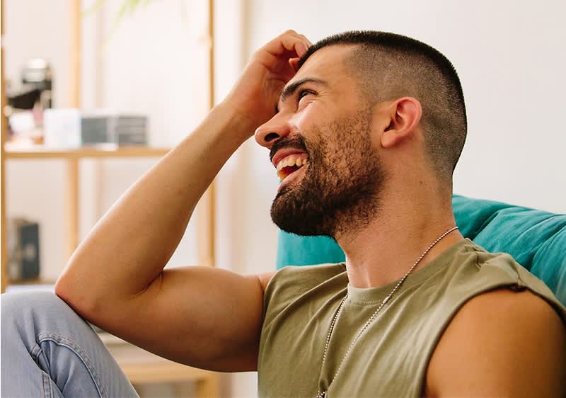 ein Mann mit kurzem Haar und Ärmellosem Olivgrünem Shirt sitzt seitlich auf einem Sofa, eine Hand hebt er an die Stirn, im Hintergrund Regalfach mit Büchern und Deko
