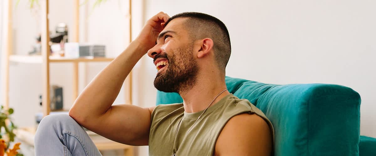ein Mann mit kurzem Haar und Ärmellosem Olivgrünem Shirt sitzt seitlich auf einem Sofa, eine Hand hebt er an die Stirn, im Hintergrund Regalfach mit Büchern und Deko