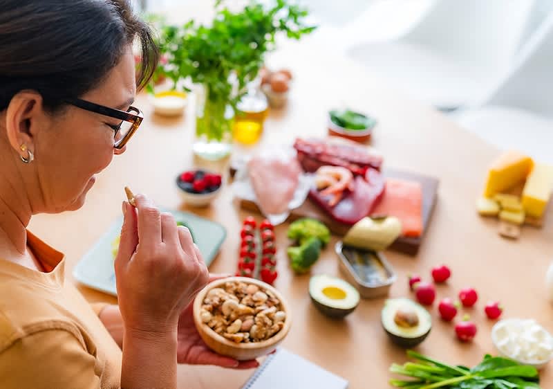 Frau von hinten links am Tisch, sie hält eine kleine Schale mit gemischten Nüssen; auf dem Tisch davor verschiedene Lebensmittel wie Avocadohälften, Eier, Beeren, Käsestücke, Aufschnitt, Gemüse und Kräuter in lockerer Anordnung