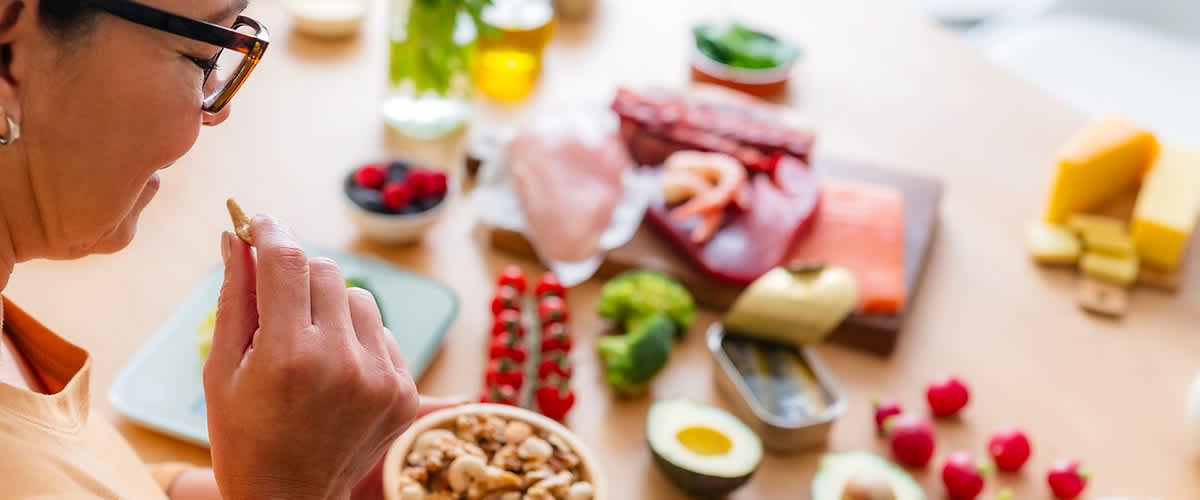 Frau von hinten links am Tisch, sie hält eine kleine Schale mit gemischten Nüssen; auf dem Tisch davor verschiedene Lebensmittel wie Avocadohälften, Eier, Beeren, Käsestücke, Aufschnitt, Gemüse und Kräuter in lockerer Anordnung