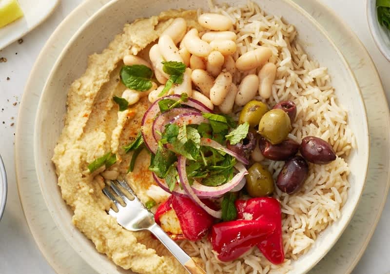 Schale mit Hummus, Reis, weißen Bohnen, grünen und schwarzen Oliven, roten eingelegten Paprika, roten Zwiebelringen und gehackten Kräutern, Gabel am Rand
