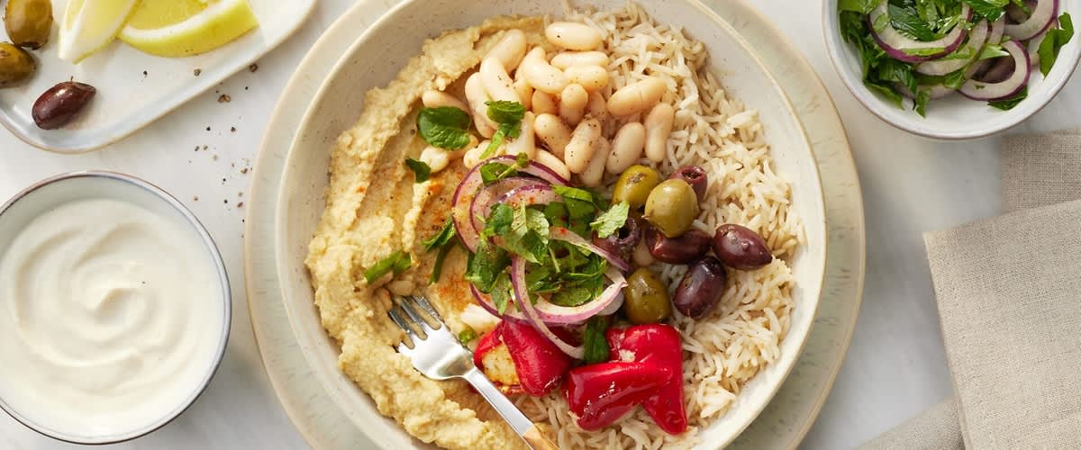 Schale mit Hummus, Reis, weißen Bohnen, grünen und schwarzen Oliven, roten eingelegten Paprika, roten Zwiebelringen und gehackten Kräutern, Gabel am Rand
