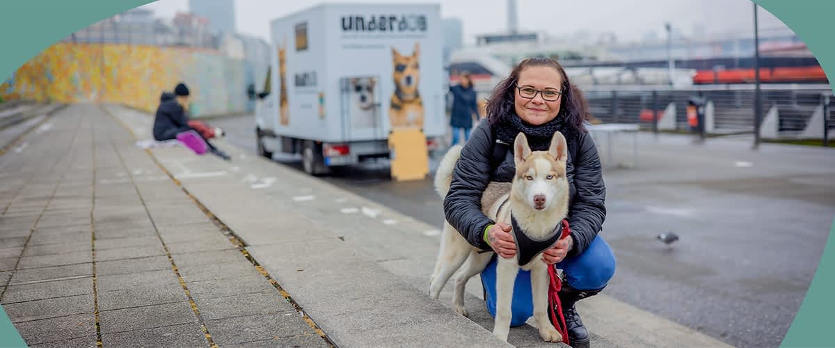 Person kniend am Ufer hält einen angeleinten Husky mit hellem Fell, im Hintergrund ein weißer Anhänger mit Hundebild und underdog Schriftzug.