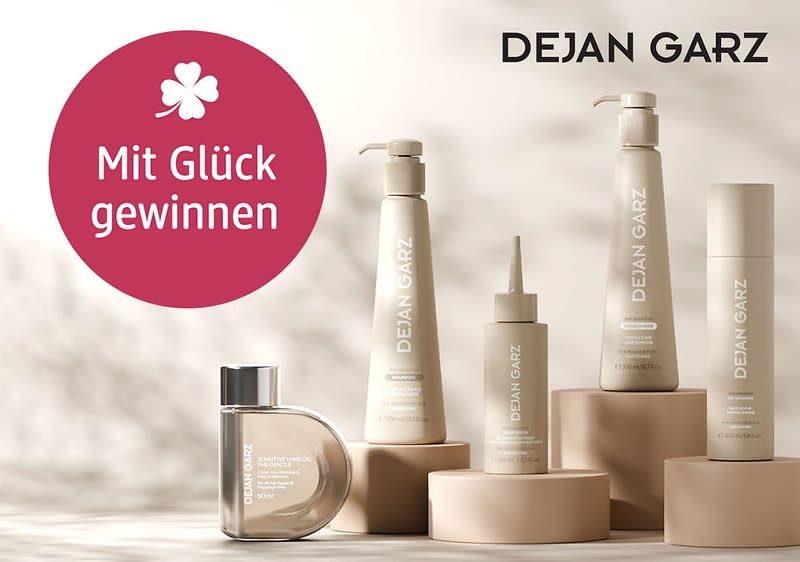 Mehrere beige-braune DEJAN GARZ Pflegeprodukte in Pump- und Flaschenform auf runden Sockeln, links ein roter Kreis mit Weißtext „Mit Glück gewinnen“ und ein Kleeblatt-Symbol