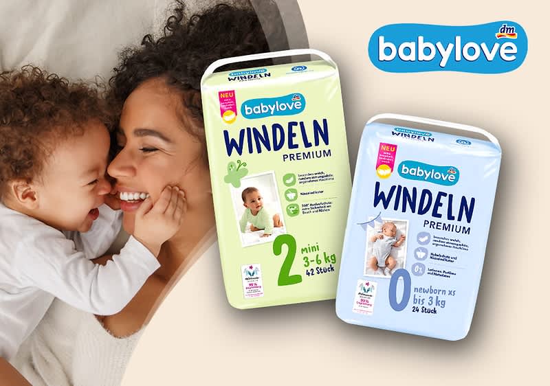 Zwei Packungen babylove Windeln Premium, links grüne Packung Größe 2 (mini 3–6 kg, 42 Stück), rechts blaue Packung Größe 0 (newborn bis 3 kg, 24 Stück), im Hintergrund Mutter lächelt über schlafendes Baby und babylove-Logo oben rechts