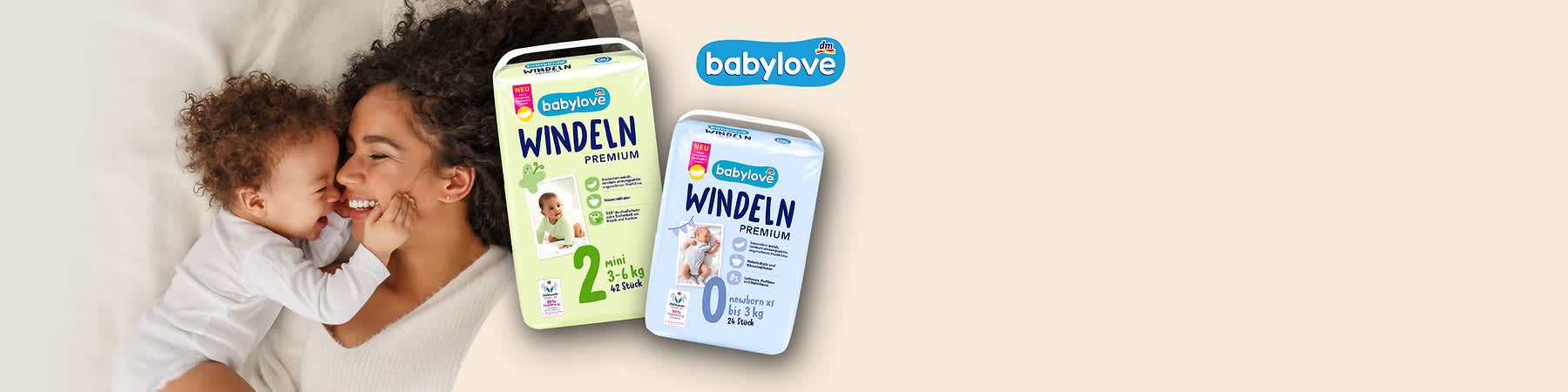 Zwei Packungen babylove Windeln Premium, links grüne Packung Größe 2 (mini 3–6 kg, 42 Stück), rechts blaue Packung Größe 0 (newborn bis 3 kg, 24 Stück), im Hintergrund Mutter lächelt über schlafendes Baby und babylove-Logo oben rechts
