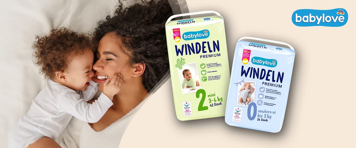 zwei Packungen babylove Windeln Premium, links grün gekennzeichnete Größe 2 „mini 3–6 kg, 42 Stück“, rechts hellblaue Packung Größe 0 „newborn xs bis 3 kg, 24 Stück“, oben rechts babylove Logo, links unscharf eine liegende erwachsene Person mit Baby (Gesichter verpixelt)