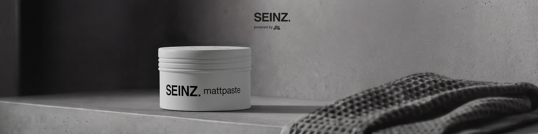Weiße runde Dose mit schwarzer Aufschrift „SEINZ. mattpaste“ auf grauem Betonuntergrund, rechts oben Logo „SEINZ. powered by dm“. Bild mithilfe von KI erstellt