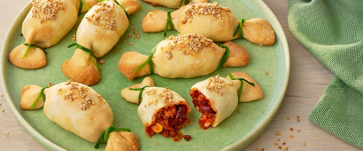 Mehrere kleine, gebackene Teigtaschen mit Sesam bestreut auf grünem Teller, eine aufgeschnittene zeigt eine Füllung aus Mais und Tomaten-/Paprikamischung, einige Teigtaschen sind mit grünen Kräuterbändern gebunden