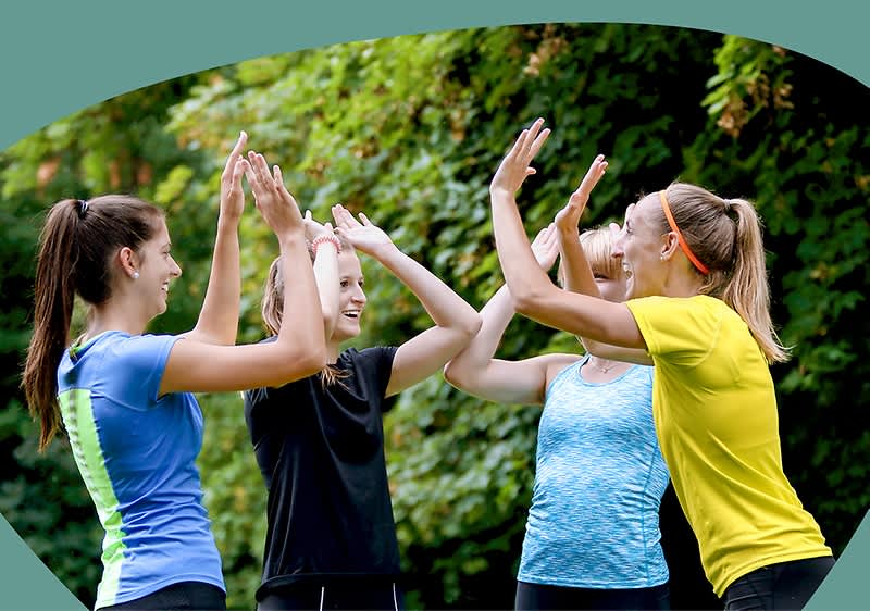 Vier junge Frauen in Sportkleidung stehen im Freien und geben sich gleichzeitig Highfives.