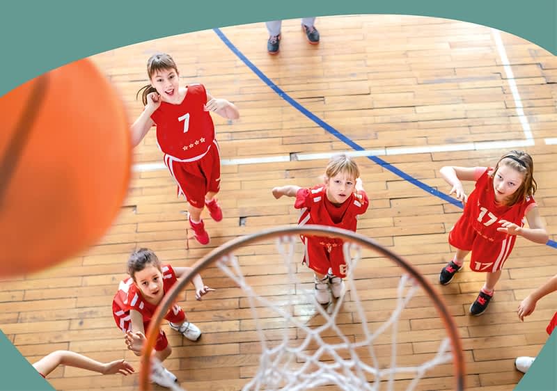 Junge Basketballspielerinnen in roten Trikots schauen von unten aus der Hallenmitte zum Korb, im Vordergrund Ball nahe am Reifen und Netz sichtbar.