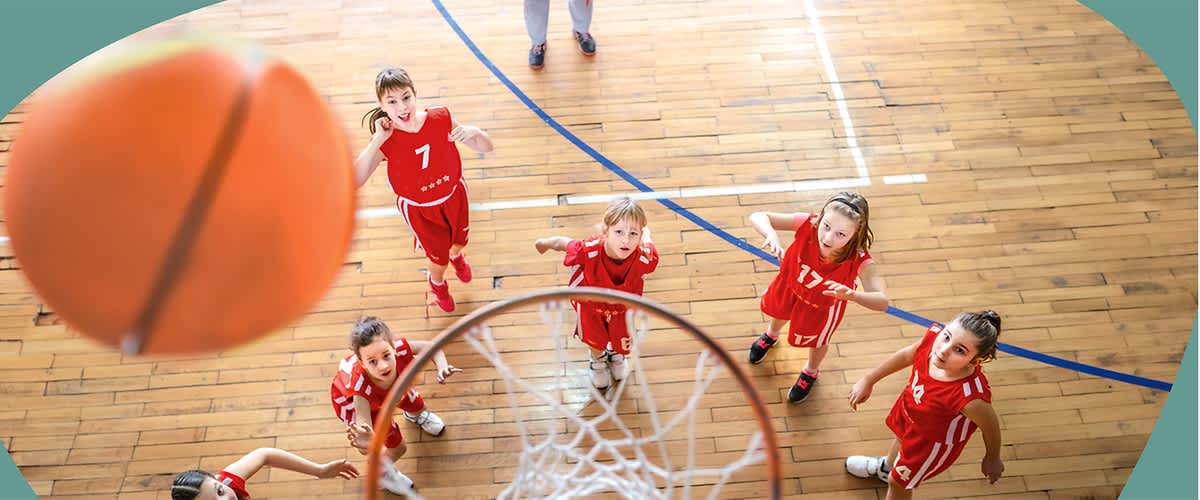 Junge Basketballspielerinnen in roten Trikots schauen von unten aus der Hallenmitte zum Korb, im Vordergrund Ball nahe am Reifen und Netz sichtbar.