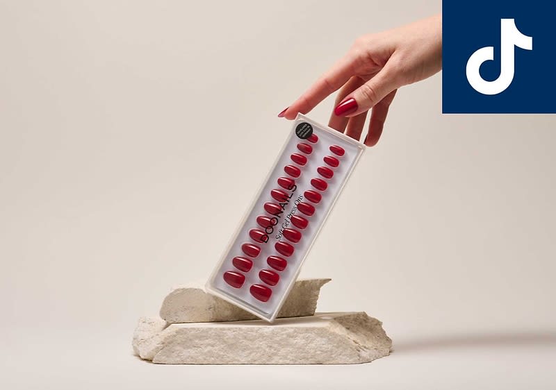 Hand hĂ€lt eine transparente Blisterpackung mit roten kĂŒnstlichen FingernĂ€geln, Packung zeigt mehrere NagelgröĂen und schwarzen Aufkleber, darunter zwei unregelmĂ€Ăige weiĂe Steinplatten als Unterlage