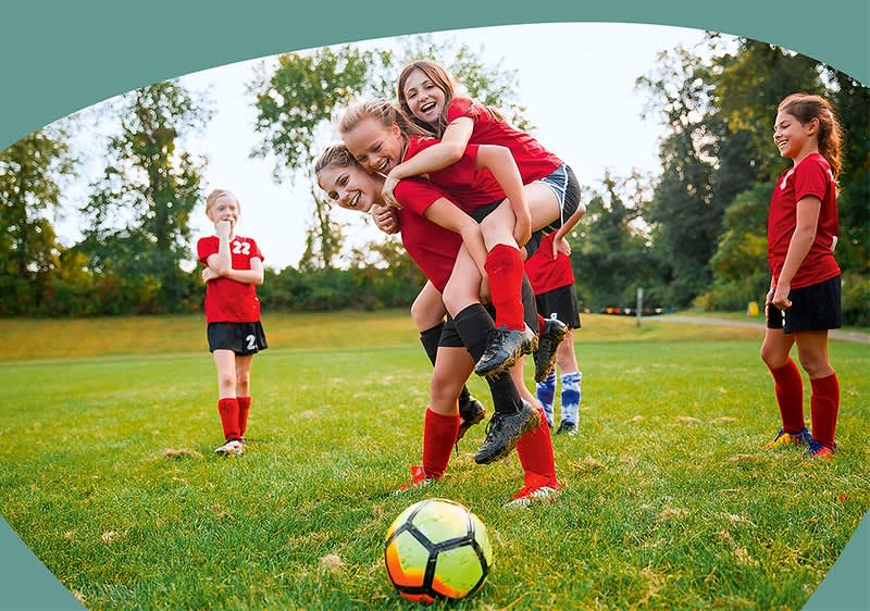 Fünf Mädchen in roten Fußballtrikots auf einem Rasen: zwei Mädchen in der Bildmitte, eine trägt die andere auf dem Rücken und beide lachen, ein Fußball liegt vorn auf dem Gras, links und rechts stehen weitere Mitspielerinnen in roten Trikots und Stutzen, Hintergrund Bäume und Parklandschaft