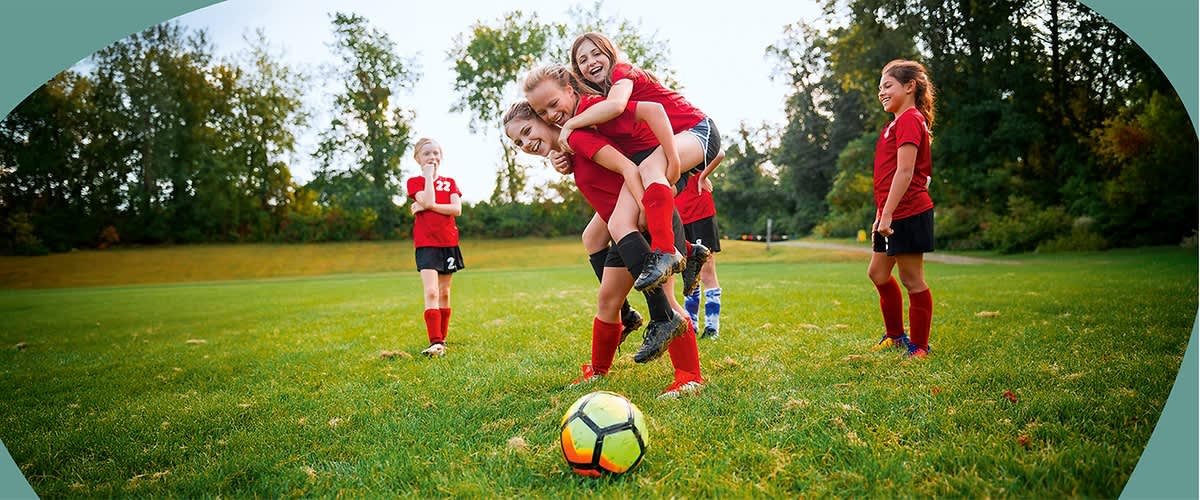 Fünf Mädchen in roten Fußballtrikots auf einem Rasen: zwei Mädchen in der Bildmitte, eine trägt die andere auf dem Rücken und beide lachen, ein Fußball liegt vorn auf dem Gras, links und rechts stehen weitere Mitspielerinnen in roten Trikots und Stutzen, Hintergrund Bäume und Parklandschaft