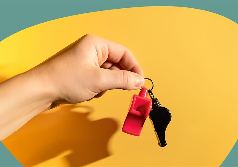 Hand hält Schlüsselring mit zwei Trillerpfeifen, eine rot und eine schwarz, vor gelbem Hintergrund
