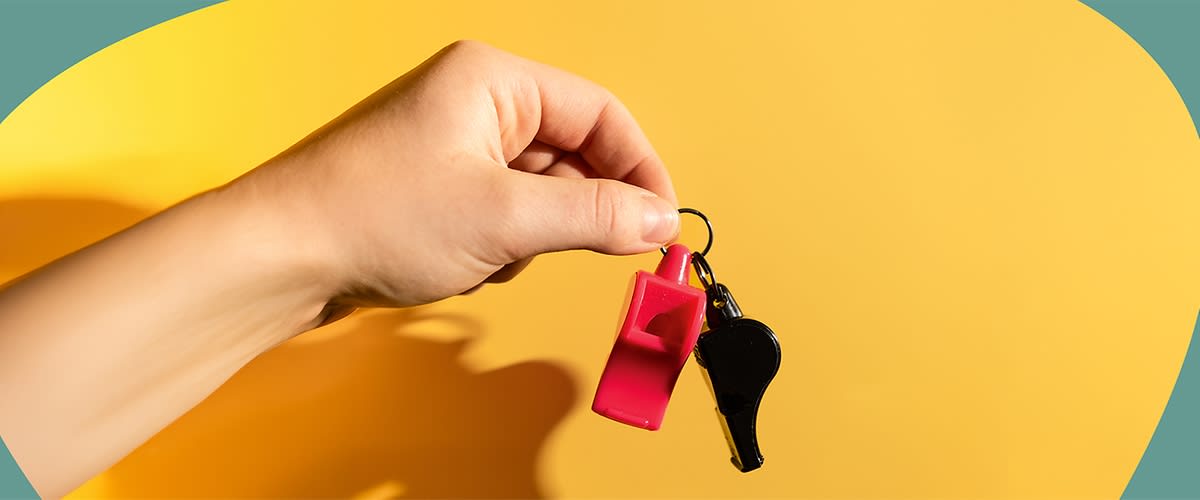 Hand hält Schlüsselring mit zwei Trillerpfeifen, eine rot und eine schwarz, vor gelbem Hintergrund