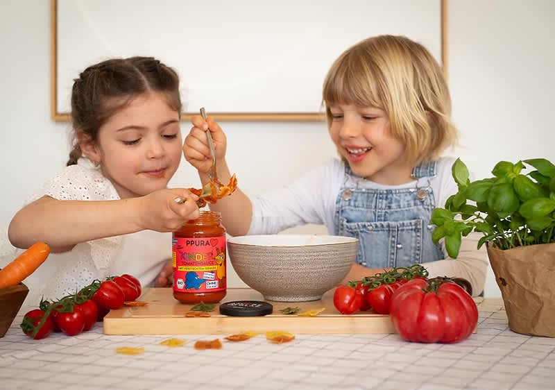 Zwei Kinder sitzen an einem Tisch und verteilen Tomatensauce aus einem Glas auf einem Löffel, auf dem Tisch liegen frische Tomaten, eine Karotte und ein Basilikumtopf, auf dem Glas ist die Aufschrift „PURA“ und „KINDER TOMATENSAUCE“ visible