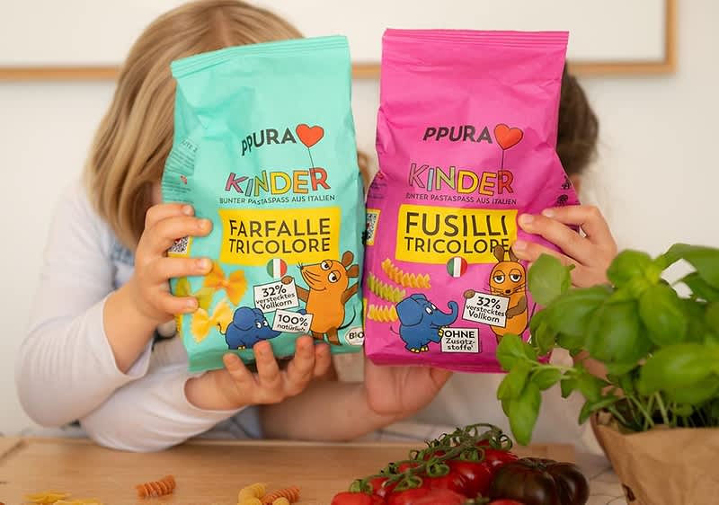 Zwei Kinderhände halten jeweils eine bunte Pastatüte der Marke „PPURA KINDER“, links türkis mit Aufschrift „Farfalle Tricolore“, rechts pink mit Aufschrift „Fusilli Tricolore“, auf den Packungen kindliche Tierillustrationen und Angaben wie „32% Vollkorn“ und „100%“ bzw. „ohne Zuckerzusatz“ sichtbar