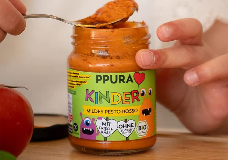 Glas mit orangefarbenem Kinder-Pesto der Marke PPURA, Aufschrift „PPURA KINDER mildes Pesto rosso“, Symbole „mit Frischkäse“, „ohne Zuckerzusatz“ und Bio-Siegel, daneben ein Löffel, mit Pesto befüllt, und Kinderhände ohne Hintergrunddetails