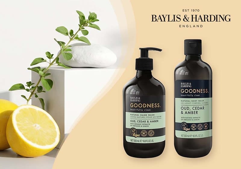 Zwei dunkelbraune Flaschen der Marke Baylis & Harding mit Aufschrift „GOODNESS. Oud, Cedar & Amber“, links eine Pumppenspender-Flasche für Handseife, rechts eine Flasche für Body Wash, links im Vordergrund halbierte Zitronen und ein grüner Zweig als Dekoration