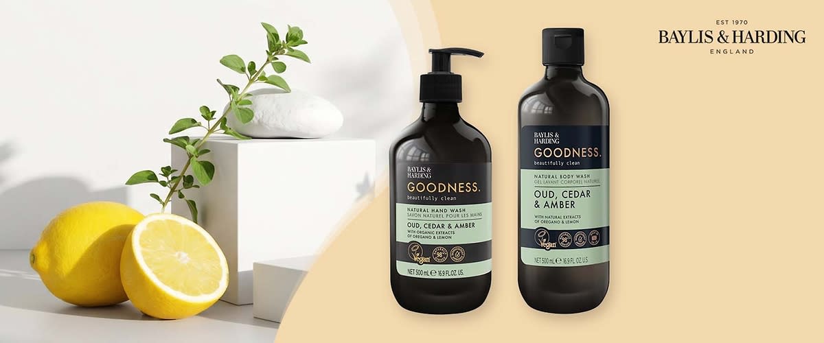 Zwei dunkelbraune Flaschen der Marke Baylis & Harding mit Aufschrift „GOODNESS. Oud, Cedar & Amber“, links eine Pumppenspender-Flasche für Handseife, rechts eine Flasche für Body Wash, links im Vordergrund halbierte Zitronen und ein grüner Zweig als Dekoration