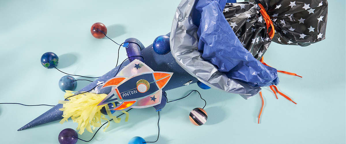 Papierkostüm und Accessoires im Weltraumdesign: eine blau bemalte Papprolle mit daran befestigter Rakete mit Aufschrift „ANTON“, gelbem Bast-Raketenschub, mehreren an Schnüren hängenden Planetenkugeln und ein silber-blaues Brust- oder Umhangteil mit Sternenmuster und orangenen Bändern
