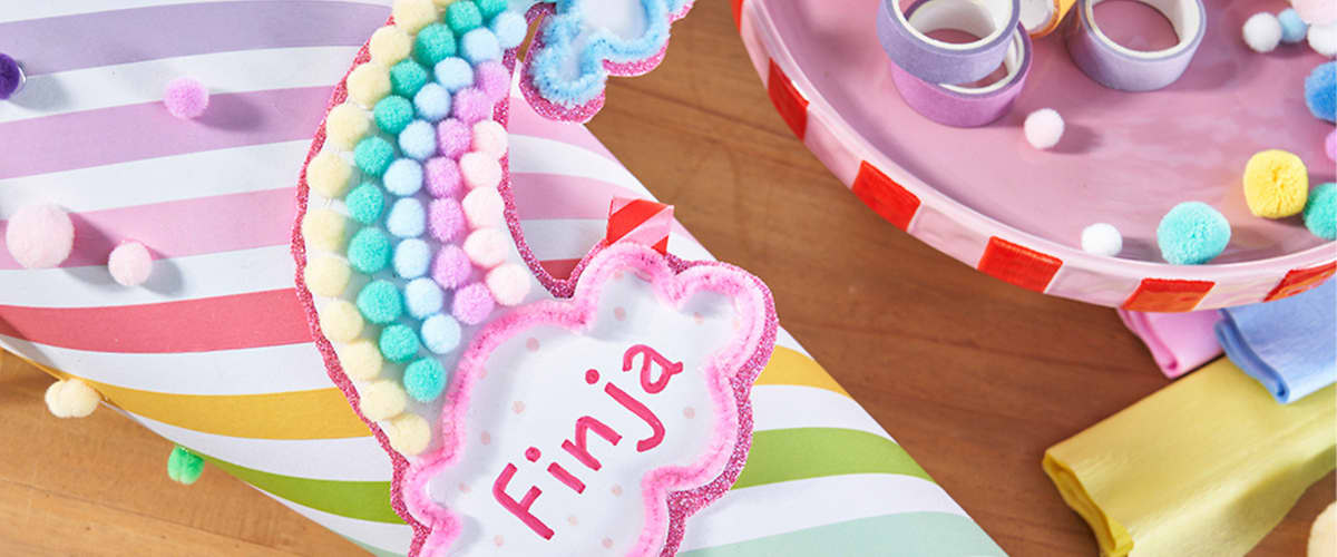 Personalisierter Namensanhänger in Wolkenform mit roter Aufschrift Finja, dekoriert mit farbigen Pompons, auf gestreiftem Geschenkpapier neben buntes Klebeband und weiteren Bastelmaterialien