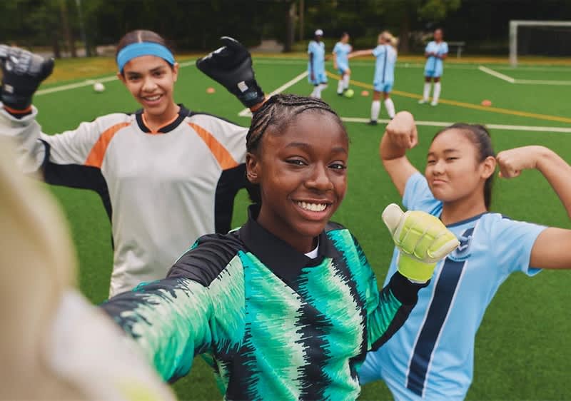 Drei Jugendfußballerinnen in Torwartausrüstung posieren jubelnd auf dem Spielfeld, vorn eine Torfrau in grün-schwarzem Trikot mit Handschuhen, rechts eine Mitspielerin in hellblauem Trikot zeigt muskelnde Armpose, im Hintergrund weitere Spielerinnen auf dem Rasen ohne erkennbare Gesichter