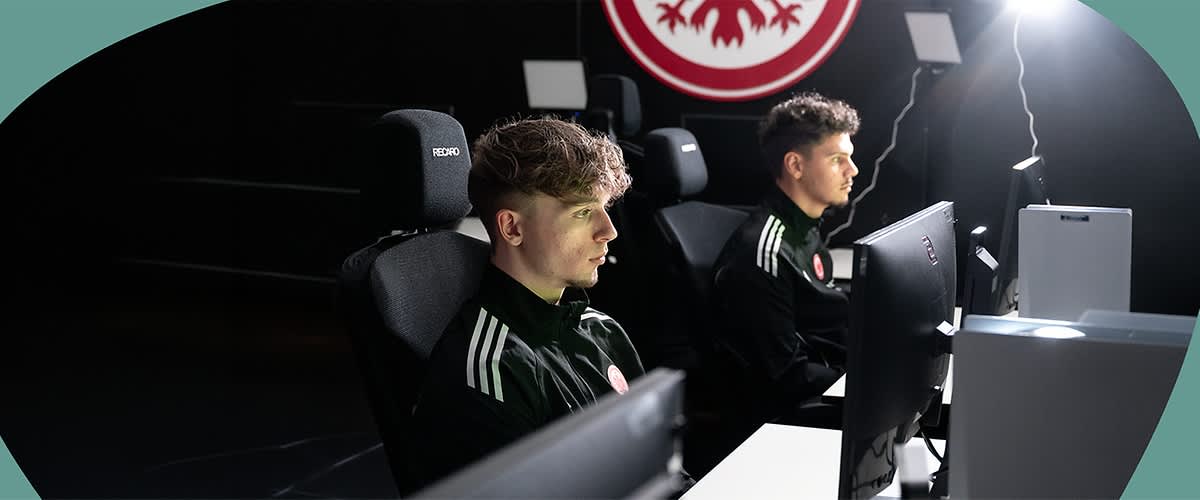 Romeo Rissling und Maxime Muscheid sitzen nebeneinander an ihren Gaming-Stationen mit Monitoren und schwarzen Recaro-Gamingstühlen, beide tragen dunkle Trainingsjacken mit weißen Schulterstreifen und einem runden Vereinslogo, im Hintergrund ein großes rundes Vereinswappen auf dunklem Hintergrund