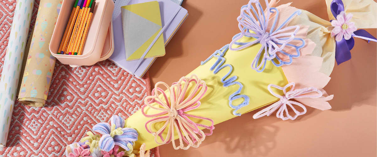 Gelbe Schultüte mit pastellfarbenen Wollblumen und Schriftzug aus Wolle, daneben Buntstifte in einem Etui, Notizbücher und ein rosafarbenes Musterläufer