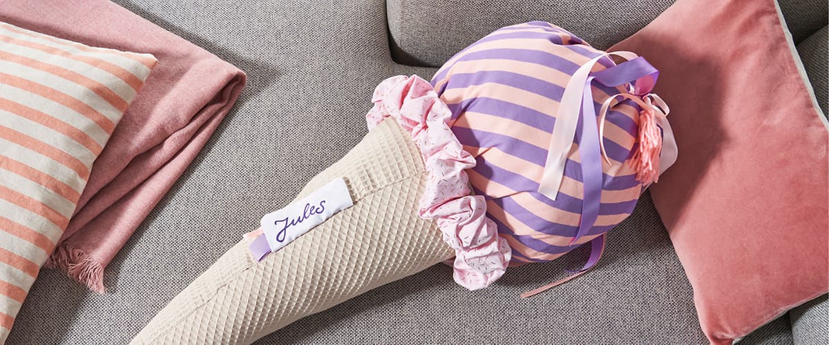 Stoff-Schultüte in Eisförmigem beige-waffeligen Köper mit aufgenähter Namenskarte „Jules“ und lila-rosa gestreiftem, rüschverziertem Tüllenabschluss mit Bändern, liegt auf einem grauen Sofa