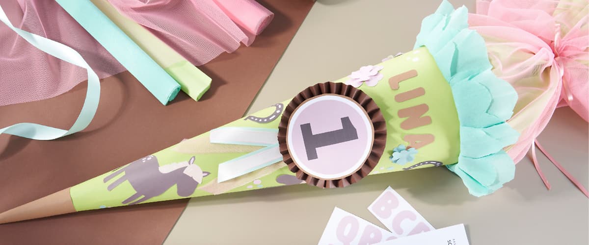 Grüne Schultüte mit blauem Papierbesatz, Zierblumen und Namensschrift „LINA“, darauf eine rosafarbene Schleife mit der Zahl 1, umgeben von Bastelmaterialien wie farbigem Papier und Aufklebern
