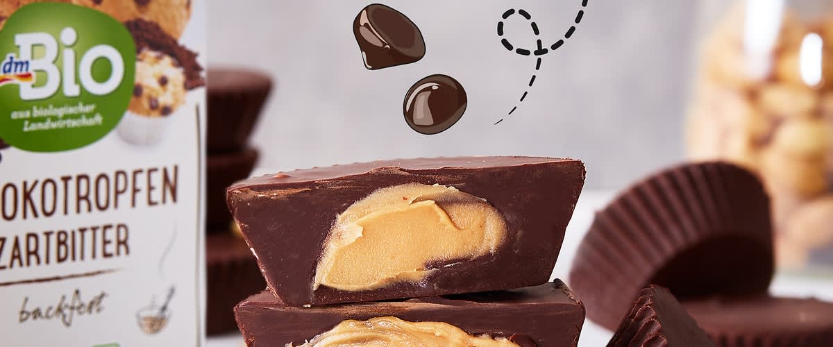 Drei schokoladenüberzogene Erdnussbutter-Täfelchen, zwei halbiert und übereinander gestapelt, im Vordergrund, daneben aufgeklappte Papierförmchen und links eine Verpackung mit Aufschrift „dmBio Zartbitter Schokotropfen“