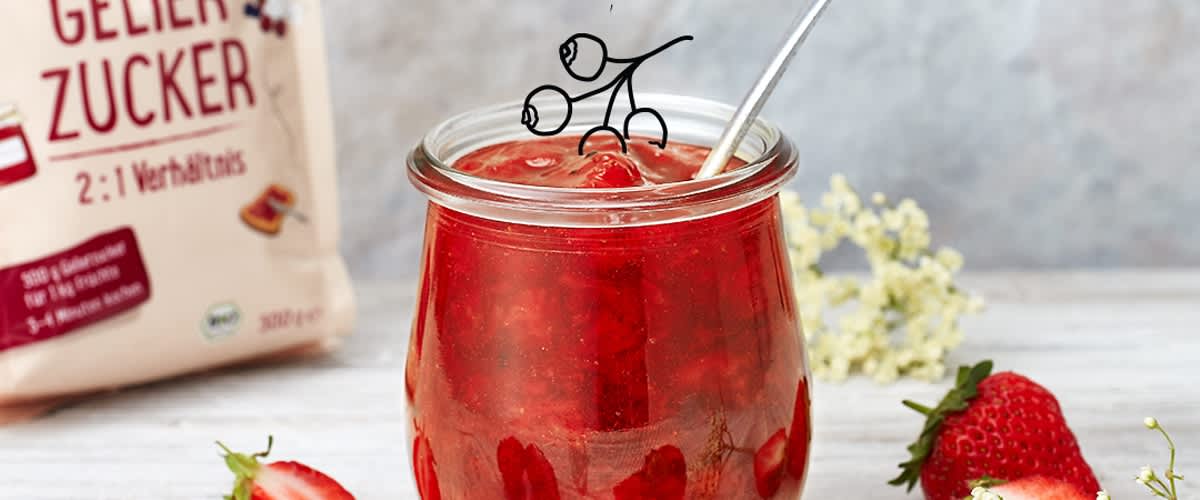 Glas mit roter Erdbeerkonfitüre und Löffel, umgeben von frischen Erdbeeren und einer Packung Bio-Gelierzucker im Hintergrund