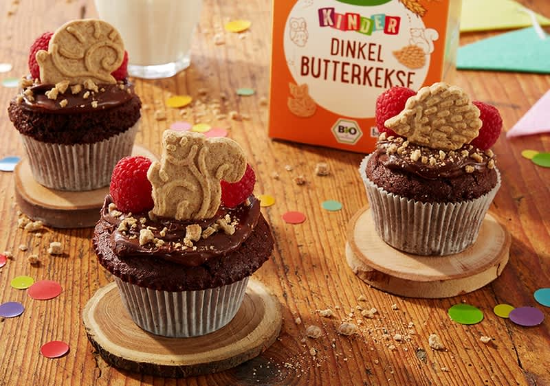 Drei Schokoladen-Cupcakes mit Schokoglasur, gehackten Nüssen, je einer Himbeere und je einem Dinkel-Butterkeks als Dekoration, im Hintergrund eine orange Verpackung mit der Aufschrift „Dinkel Butterkekse“
