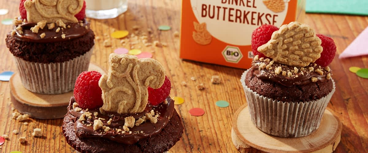 Drei Schokoladen-Cupcakes mit Schokoglasur, gehackten Nüssen, je einer Himbeere und je einem Dinkel-Butterkeks als Dekoration, im Hintergrund eine orange Verpackung mit der Aufschrift „Dinkel Butterkekse“