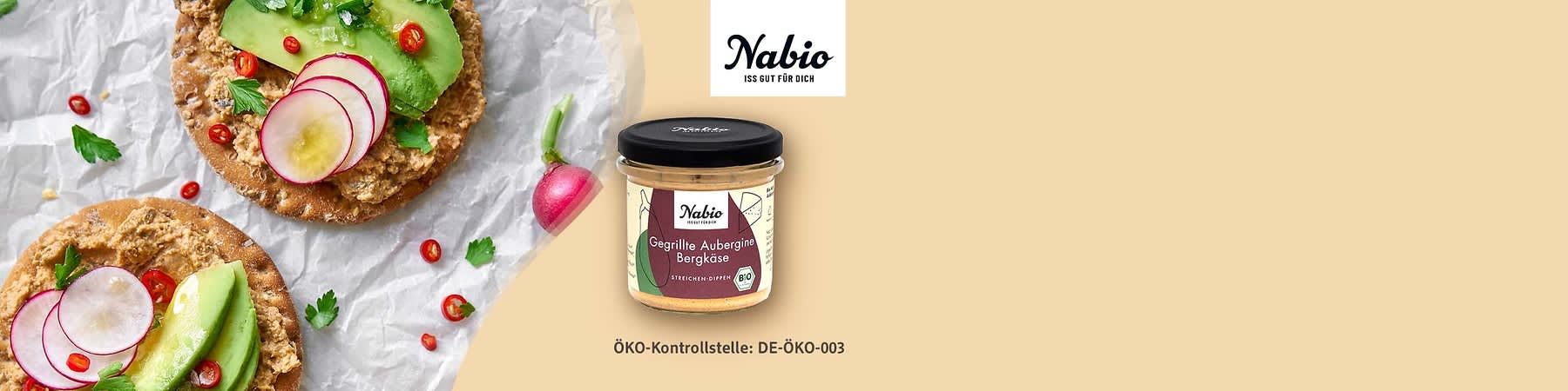 Glas mit Nabio Aufschrift und Etikett „Gegrillte Aubergine Bergkäse“, daneben zwei Cracker mit Aufstrich, Avocadoscheiben, Radieschenscheiben, Petersilie und roten Chiliringeln, grüner Hintergrund und Schriftzug „Nabio“