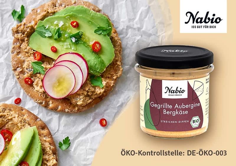 Glas mit Nabio Aufschrift und Etikett „Gegrillte Aubergine Bergkäse“, daneben zwei Cracker mit Aufstrich, Avocadoscheiben, Radieschenscheiben, Petersilie und roten Chiliringeln, grüner Hintergrund und Schriftzug „Nabio“