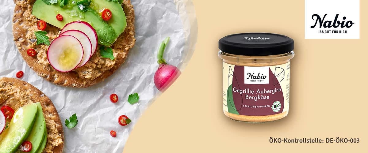 Glas mit Nabio Aufschrift und Etikett „Gegrillte Aubergine Bergkäse“, daneben zwei Cracker mit Aufstrich, Avocadoscheiben, Radieschenscheiben, Petersilie und roten Chiliringeln, grüner Hintergrund und Schriftzug „Nabio“