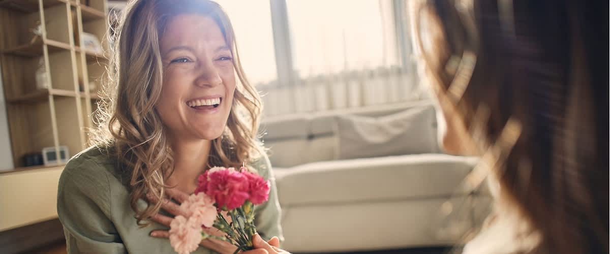 Frau sitzt im Wohnzimmer und lächelt, während sie von einer anderen Person einen kleinen Blumenstrauß mit rosa und pinken Nelken entgegenimmt