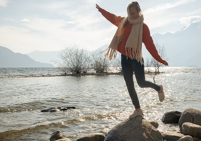 Frau mit Schal und rotem Pullover balanciert auf einem Felsen am Ufer eines Sees, im Hintergrund sind Wasser und Berge zu sehen.