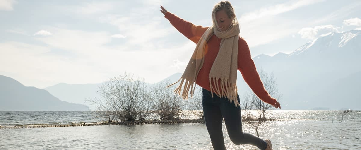 Frau mit Schal und rotem Pullover balanciert auf einem Felsen am Ufer eines Sees, im Hintergrund sind Wasser und Berge zu sehen.
