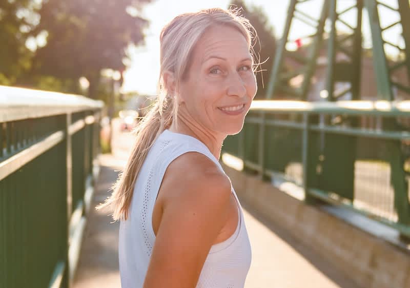 Frau im weißen ärmellosen Sporttop steht auf einer Fußgängerbrücke, leicht zur Seite gewandt, Zopf, Sonnenlicht von hinten