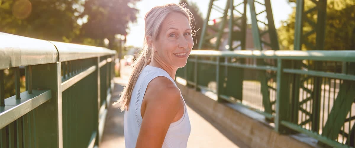 Frau im weißen ärmellosen Sporttop steht auf einer Fußgängerbrücke, leicht zur Seite gewandt, Zopf, Sonnenlicht von hinten